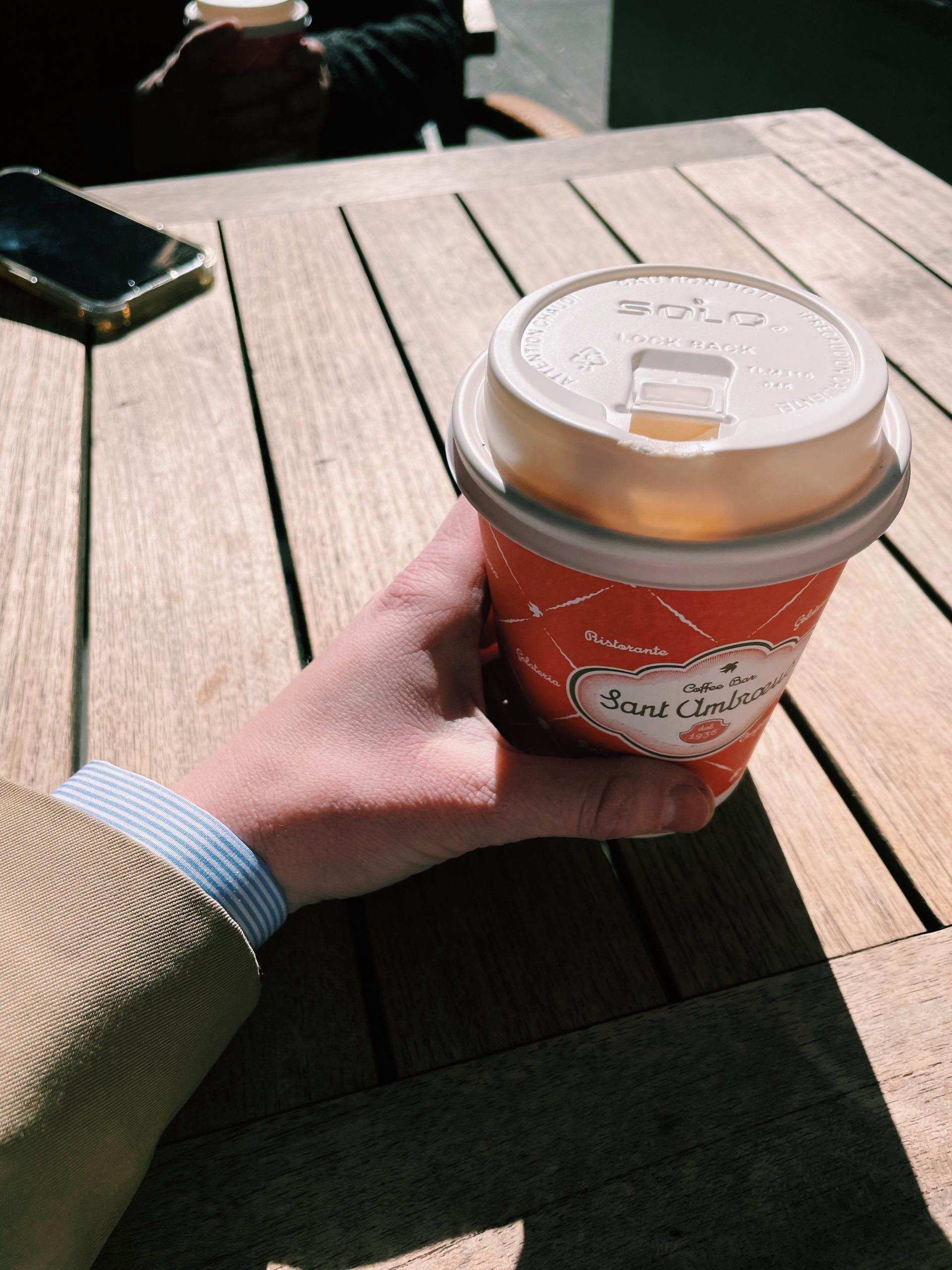 Hand holding a coffee cup from Sant Ambroeus with a blue striped popover sleeve and trench coat sleeve showing.